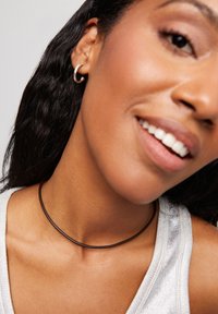 Un collar choker negro descansa sobre un cuello, con un diseño suave y minimalista. El modelo lleva una blusa sin mangas de plata, mostrando un tono de piel claro.