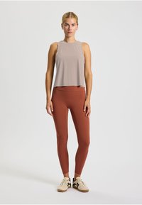 Top senza maniche grigio chiaro abbinato a leggings color ruggine. Entrambi i capi presentano un design liscio e aderente. La modella indossa sneakers beige.