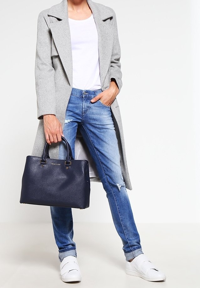 Personne portant un manteau gris clair, une chemise blanche, un jean bleu déchiré, des baskets blanches, tenant un grand sac à main bleu foncé par la poignée.