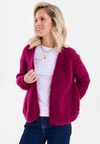 Kobieta o blond włosach ubrana w puszysty magentowy kardigan na białej koszuli i niebieskich dżinsach, uśmiechająca się i patrząca w lewo.