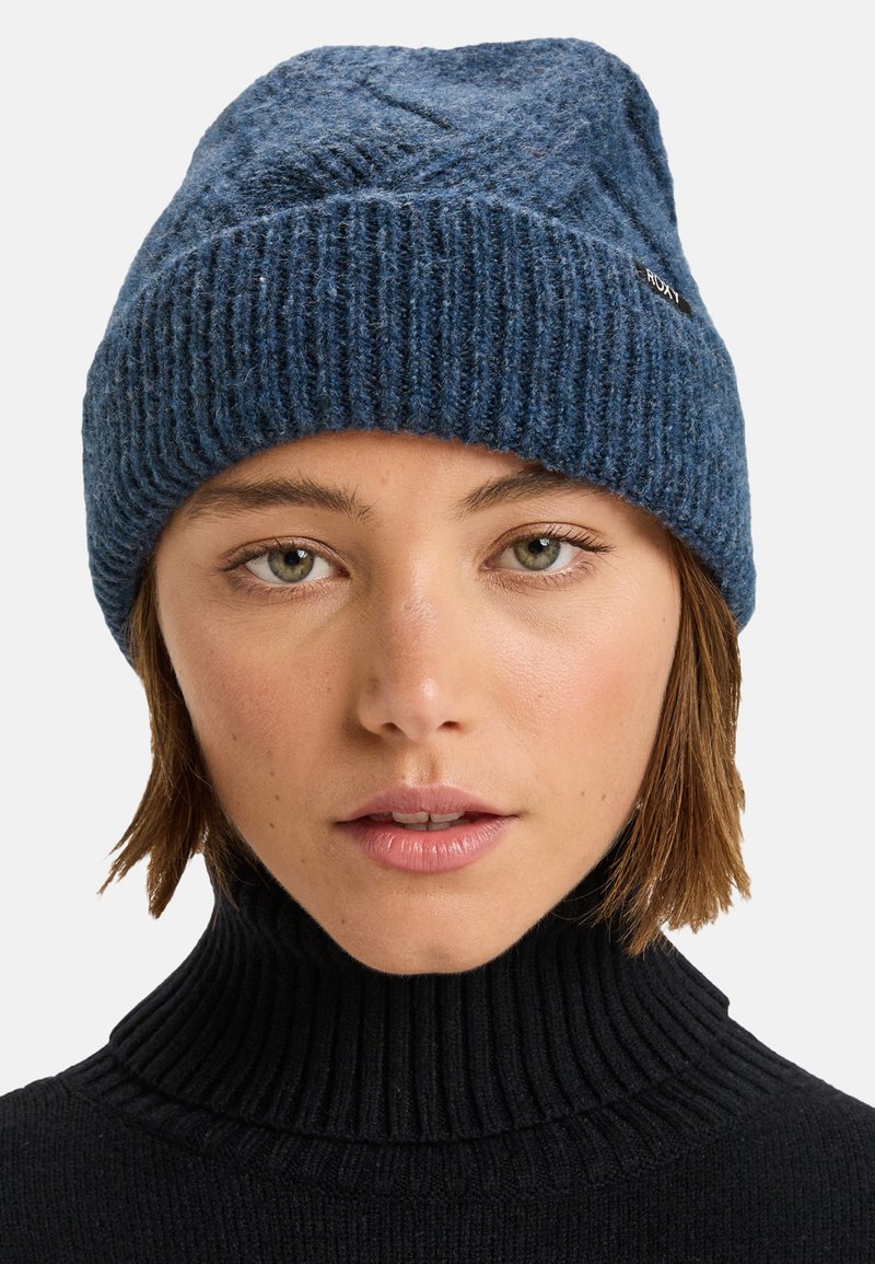 Blå strikbeanie med ribbet bund, struktureret top og subtilt mønsterdesign. Har et lille mærke på siden.