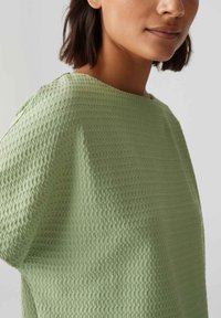 Haut vert clair en tissu texturé, présentant un col large et des manches courtes, avec un subtil motif de grille sur l'ensemble.