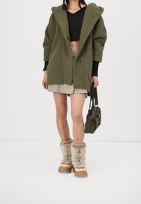 Abrigo de felpa verde con capucha grande, top negro corto, falda gris claro y botas beiges forradas de piel con suelas gruesas. Bolso verde oliva.