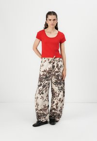 Haut rouge à manches courtes associé à un pantalon large beige avec un motif abstrait marron. Le modèle porte des chaussures noires et de grandes boucles d'oreilles en anneau.