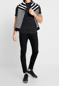 T-shirt noir sous un pull sans manches noir et blanc à rayures, associé à un jean slim noir et des chaussures de sport noires.