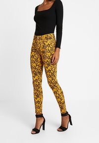 Pantalones ajustados de estampado de serpiente amarilla con una camiseta negra de canalé. Los pantalones tienen una cintura alta, textura suave de satén y sutiles acentos negros en el dobladillo.