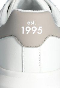 Sneaker bianco con una superficie in pelle liscia e dettagli grigio chiaro. Presenta una cucitura decorativa e il testo "est. 1995" in bianco.