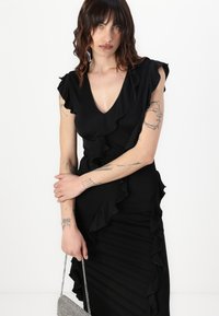 Black dress with ruffled edges, V-neck design, fitted silhouette, and side draping. Silver chain bag accentuates the look.