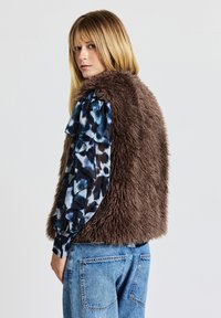 Braune Weste aus Kunstfell mit strukturierter Oberfläche, kombiniert mit einer blauen Bluse mit abstraktem Muster und Rüschenärmeln sowie hellen Denim-Jeans.