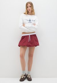 Joven mujer con los brazos cruzados, vistiendo un sudadera blanca de Nueva York, falda roja con cordón, calcetines grises y zapatillas marrones, de pie contra una pared blanca.