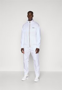 White athletic jacket with a high collar and zip closure, paired with matching pants and white sneakers, featuring a black logo detail.