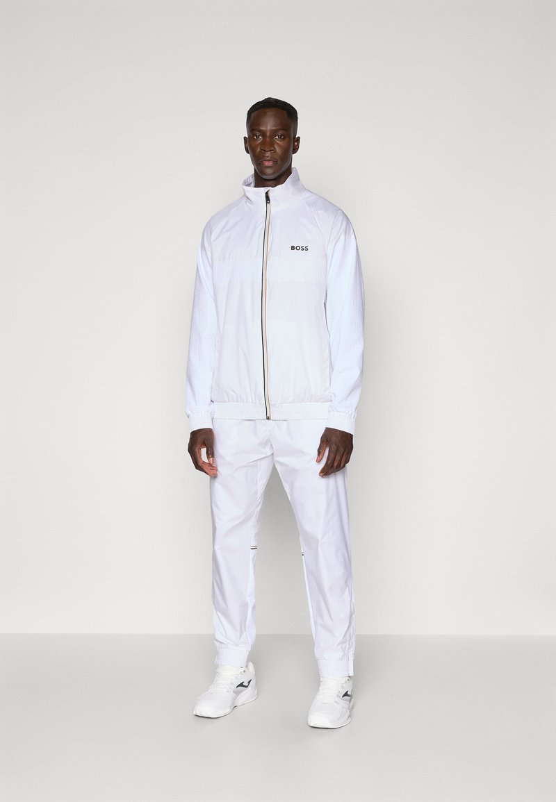 White athletic jacket with a high collar and zip closure, paired with matching pants and white sneakers, featuring a black logo detail.