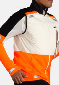 Veste rembourrée avec un col haut, présentant des panneaux blancs, noirs et orange vif, des accents réfléchissants et des poches zippées sur les côtés.