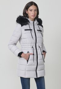 Parka acolchada de color gris claro con capucha de pelo sintético negro, cremalleras negras, cordones y bolsillos laterales. Presenta una silueta ajustada y puños acanalados.