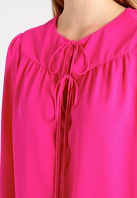 Blusa fucsia con collo arrotondato e lacci, dettagli plissettati, maniche lunghe e una texture di tessuto liscia e leggera.
