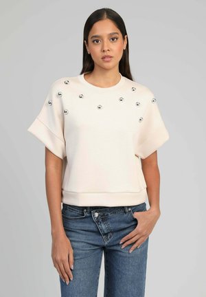 Femme portant un pull crème à manches courtes avec des clous métalliques sur la poitrine, associé à un jean bleu, debout devant un fond gris.