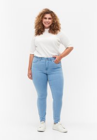 Vaqueros azul claro, ajustados, con una cintura de media altura, hechos de denim. Combinados con una camiseta blanca de manga corta y canalé y zapatillas blancas.