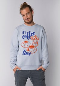 Sweatshirt bleu clair avec des graphismes sur le thème du café en orange et bleu, y compris une cafetière et des tasses, avec le texte "c'est l'heure du café."