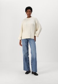 Mujer con un suéter blanco de punto trenzado, jeans de pierna ancha azules, zapatos negros y pendientes de aro, de pie contra un fondo blanco liso.