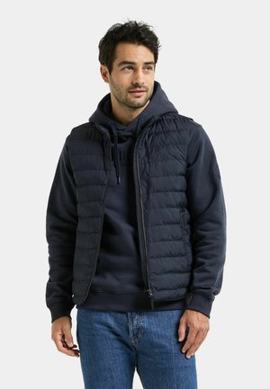 Man met een marineblauwe hoodie, marineblauw gewatteerd vest en blauwe jeans staat naar voren gericht, kijkt iets opzij, met een neutrale gezichtsuitdrukking.