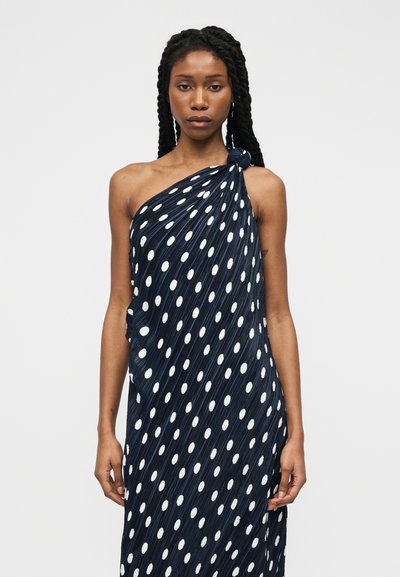 Γυναίκα με μακριά πλεκτή κότσο, φορεμένη με μπλε σκούρο φόρεμα ενός ώμου με λευκά πουά, σε μονόχρωμο φόντο.