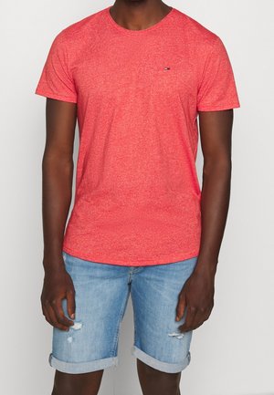 Homme portant un t-shirt rouge chiné à manches courtes et un short en jean bleu clair déchiré avec des ourlets retroussés sur un fond uni.