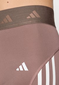 Lys lilla treningstights har en bred brun midjebånd med utskårne geometriske detaljer og tre hvite striper langs siden.