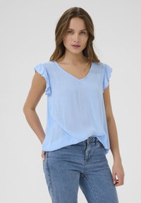 Haut bleu clair à manches courtes avec des accents en volants, encolure en V et texture douce. Poches soulignées à l'avant avec une coupe décontractée.