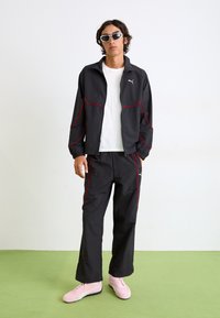 Zwarte trackjack en broek met rode accenten. Licht materiaal met een relaxte pasvorm. Gemodeerd met een wit shirt en roze sneakers.