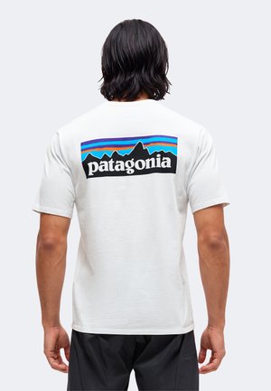 Uomo con capelli scuri fino alle spalle che indossa una t-shirt bianca Patagonia con un logo colorato di montagna sulla schiena, abbinata a pantaloni neri.
