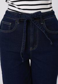 Σκούρο μπλε τζιν παντελόνι με δέσιμο στη μέση, με μπροστινές τσέπες και ραφές αντίθετου χρώματος. Το ύφασμα φαίνεται ανθεκτικό με λεία υφή.