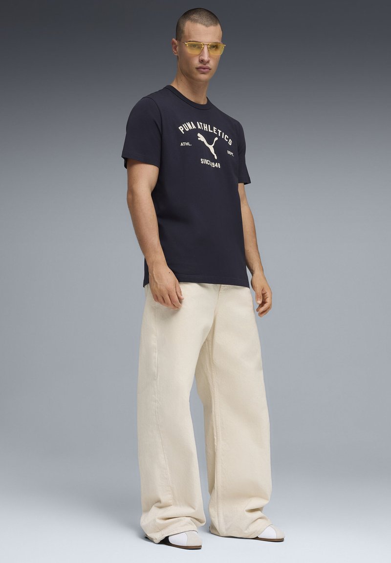 Navy t-shirt with white Puma logo, relaxed cream wide-leg pants, and yellow sunglasses. Smooth fabric textures and minimalistic design accents.
