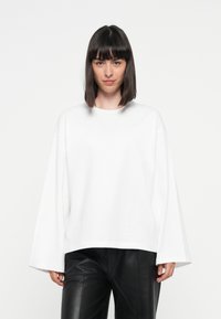 White oversized long-sleeve top with a round neck, made of soft fabric, paired with black faux leather pants, displayed against a neutral background.