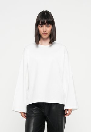 White oversized long-sleeve top with a round neck, made of soft fabric, paired with black faux leather pants, displayed against a neutral background.