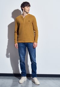 Muž stojící uvnitř ve žlutém dlouhém tričku, modrých džínách a bílých teniskách, dívající se vlevo proti bílé zdi.