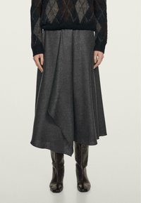 Gonna midi grigia in lana con orlo asimmetrico, texture liscia e caduta morbida. Indossata con un maglione nero a fantasia e stivali sopra al ginocchio.