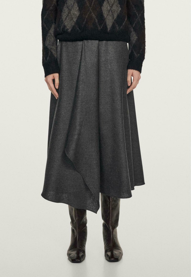 Gonna midi grigia in lana con orlo asimmetrico, texture liscia e caduta morbida. Indossata con un maglione nero a fantasia e stivali sopra al ginocchio.