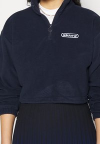 Person som bär en marinblå Adidas tröja med fyrdelad dragkedja och logotyp på bröstet, tillsammans med en plisserad mörk kjol, accessoriserad med ett silverfärgat kedjearmband.