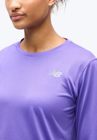 Κοντινή λήψη ενός ατόμου που φοράει ένα μωβ αθλητικό μπλουζάκι με υφή και μακριά μανίκια, με λευκό λογότυπο New Balance στο στήθος.