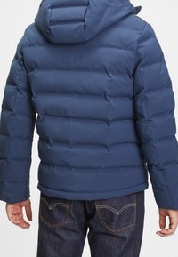 Tmavě modrá prošívaná bunda s kapucí a elastickými manžetami, doplněná tmavými džínami s kontrastním prošíváním.