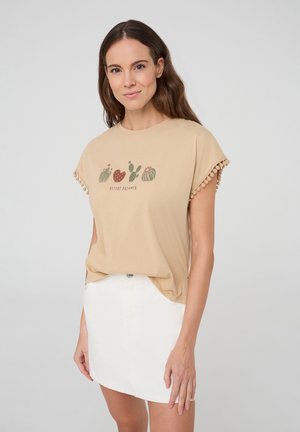 T-shirt beige a maniche corte con rifiniture in pompon, caratterizzato da una grafica di cactus e la scritta "DESERT DREAMER". Abbinato a una gonna di denim bianca.