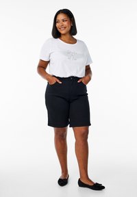 Camiseta blanca de manga corta con texto impreso y detalles gráficos, combinada con pantalones cortos de mezclilla negra de talle alto y zapatos planos negros.