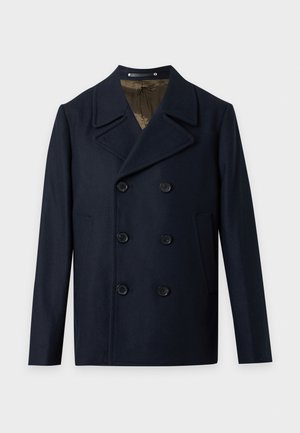 Un homme porte un manteau croisé bleu marine, un pantalon noir et des baskets rouges sur un fond clair uni.