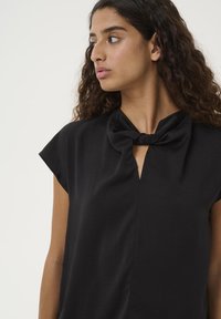 Donna con capelli ricci scuri che indossa un top nero con un dettaglio a fiocco vicino al collo, che guarda verso sinistra su uno sfondo chiaro e semplice.