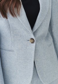 Světlomodrý herringbone blazer s klopami, jednotlivým hnědým zapínáním na knoflík a decentními bočními kapsami. Viditelné detaily texturované tkaniny.