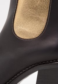 Bottine en cuir noir avec une insertion de tissu texturé doré, bout rond, coutures détaillées le long du bord et talon épais.