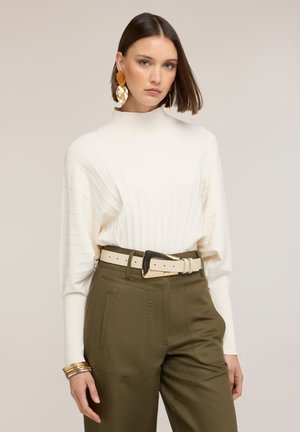 Jonge vrouw die een witte geribbelde trui draagt, olijfgroene hoge taillebroek met een crèmekleurige riem, grote gouden oorbellen en gouden armbanden.