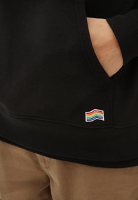 Hand in der Tasche eines schwarzen Hoodies mit kleinem, regenbogenfarbenem Flaggenaufnäher nahe dem Saum, getragen über beigen Hosen.