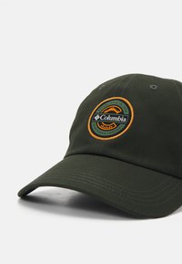 Grön basebollkeps med en böjd skärm, prydd med en broderad logotyp i orange och svart samt kontrasterande sömmar.