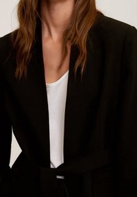 Sort blazer med en glat tekstur, åben front og et bundet bælte. Hvid skjorte under, der skaber en farvekombination.